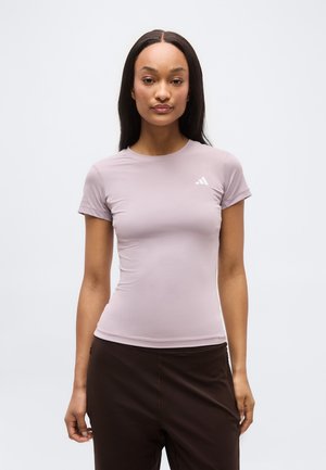 Γυναίκα με μακριά σκουρόχρωμα μαλλιά φοράει στενή αθλητική κοντομάνικη μπλούζα σε ανοιχτό μωβ χρώμα με μικρό λευκό λογότυπο και μαύρο παντελόνι, στέκεται μπροστά σε απλό φόντο.