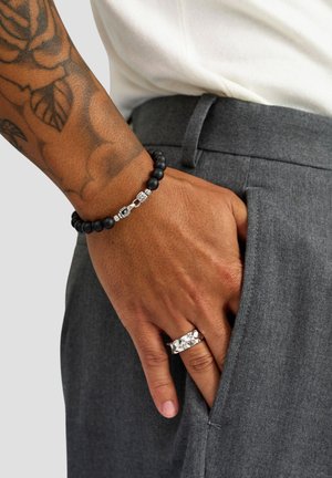 Sort perlearmbånd med sølvdetaljer på et håndled; grå bukselomme synlig; tekstureret hud og tatovering i baggrunden.