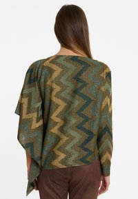 Poncho à motifs zigzag marron et vert avec des fils métalliques, présentant un large col et des manches amples, porté sur un pantalon marron.