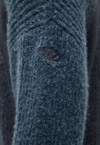 Dunkelblauer Strickpullover mit einer strukturierten, flauschigen Oberfläche und einem subtilen Chevron-Muster, versehen mit einem kleinen Lederlogo-Patch am Ärmel.