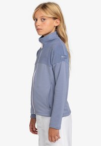 Niebieska kurtka zapinana na zamek błyskawiczny z podniesioną teksturą, wysokim kołnierzem i kontrastowym białym zamkiem. Posiada metkę marki Roxy na ramieniu.