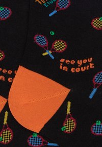 Calzini neri con motivi colorati di racchette da tennis, con accenti arancioni sul tallone e sulle punte, e testo "ci vediamo in campo" in un vivace arancione.
