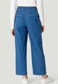 Pantalon en denim à jambes larges bleu, avec taille haute, deux poches arrière et un ourlet net. Associé à des baskets blanches et un haut à manches longues.