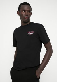 Fekete pamut póló, bal mellen egy kis rózsaszín logóval, rövid ujjal, szabványos kerek nyakkal és laza fazonnal.