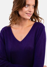 Frau mit heller Haut und welligem braunem Haar, die einen lila V-Ausschnitt-Pullover trägt, nach rechts blickt und leicht lächelt.