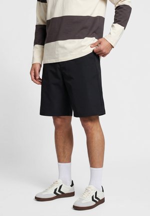 Person iført sorte shorts, hvid langærmet trøje med mørke horisontale striber, hvide sokker og hvide sneakers med sorte chevron-detaljer.