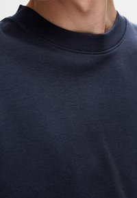 Laivansinisestä puuvillapaidasta, jossa on korkea, ribattu kaula-aukko ja sileä pinta, ja joka esittelee lähikuvan kankaan ja ompelun yksityiskohdista.