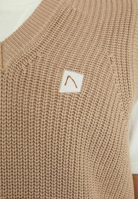 Chaleco de punto beige con escote en V y un patrón texturizado, que presenta una pequeña etiqueta blanca con un logo marrón en la parte delantera.