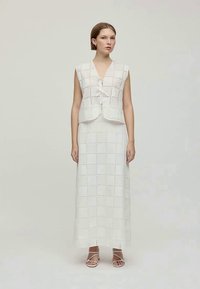 Conjunto branco de duas peças composto por uma blusa sem mangas, com botões e laço, e uma saia longa com um padrão perfurado em quadrados.