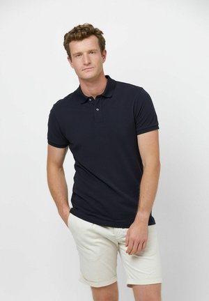 Polo a maniche corte blu navy con colletto e patta con tre bottoni, abbinata a pantaloncini di colore chiaro. Texture liscia, design aderente.