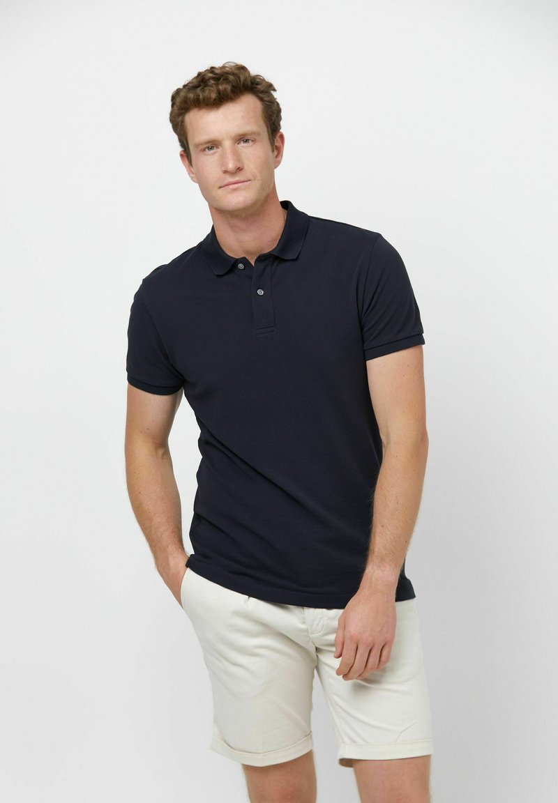 Kurzärmeliges, marineblaues Poloshirt mit Kragen und dreiknöpfigem Verschluss, kombiniert mit hellfarbenen Shorts. Glatte Textur, figurbetontes Design.