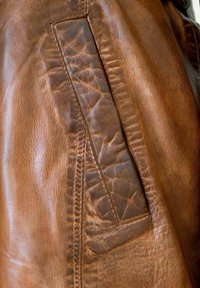 Braune Lederjacke mit strukturiertem Finish, sichtbaren Nähten und einer aufgesetzten Tasche an der Seite, die ein abgenutztes Aussehen zeigt.