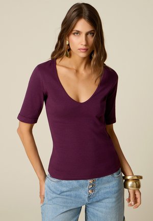 Femme portant un haut violet à col en V à manches courtes et un jean bleu clair avec des détails de boutons, accessoirisée avec des boucles d'oreilles en or et des bracelets.