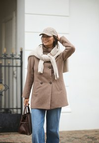 Cappotto di lana marrone doppiopetto con colletto alto, abbinato a una sciarpa di maglia color crema, jeans azzurri chiari e un cappello beige. Borsa marrone.