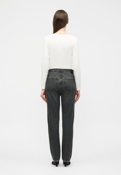 Woman seen from behind wearing a white long-sleeve top, high-waisted dark gray jeans, and black heeled shoes on a plain background.