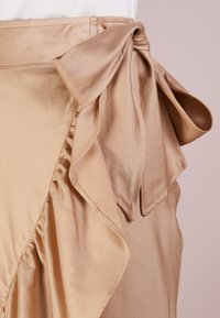 Beige hedvábná sukně s volánkovým designem a velkou mašlí v pase, s hladkou texturou a elegantním drapováním.