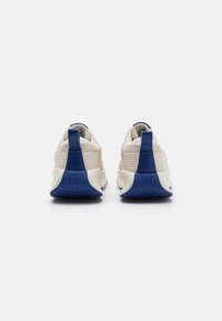 Un par de zapatillas Nike beige con pestañas y suelas azules, mostradas desde atrás sobre un fondo blanco.