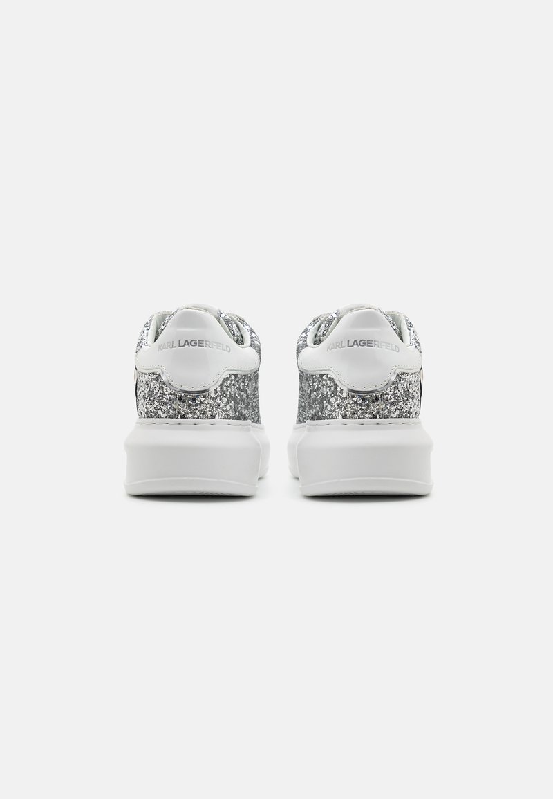 Zapatillas de deporte plateadas brillantes con una suela de goma blanca, que presentan un acabado liso y acentos de marca en el talón. Vista trasera.