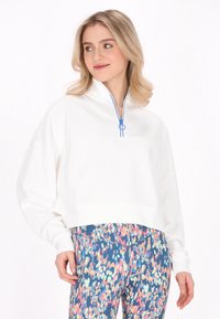 Pullover bianco accorciato con collo a zip, dotato di dettagli blu nella cerniera. Abbinato a leggings colorati e a fantasia in sfumature di blu, rosa e verde.