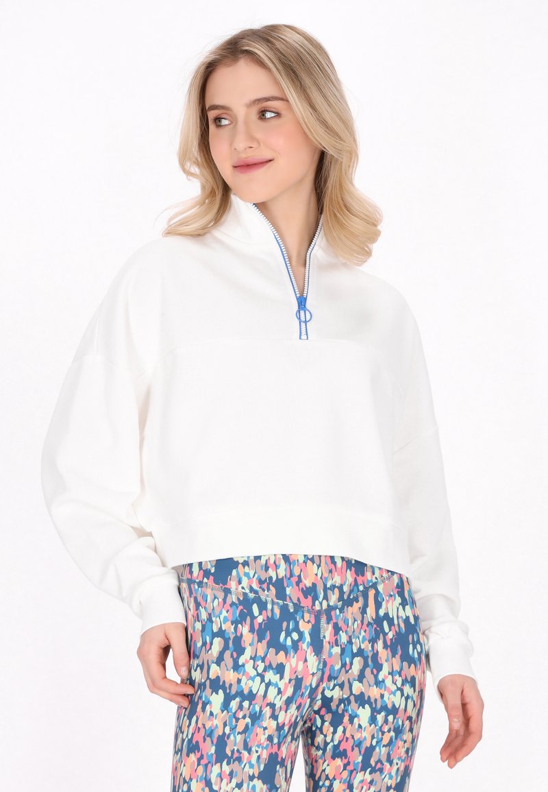 Pullover bianco accorciato con collo a zip, dotato di dettagli blu nella cerniera. Abbinato a leggings colorati e a fantasia in sfumature di blu, rosa e verde.