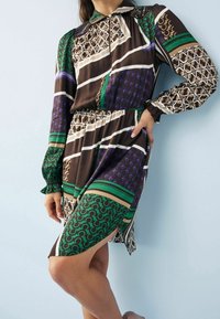 Mujer con un vestido de manga larga y hasta las rodillas, con patrones geométricos en verde, púrpura, marrón, beige y negro, de pie frente a una pared clara.