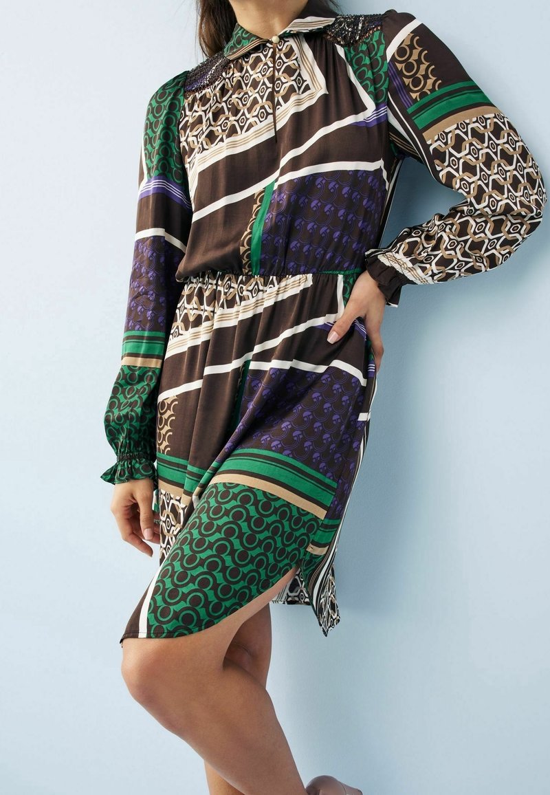 Mujer con un vestido de manga larga y hasta las rodillas, con patrones geométricos en verde, púrpura, marrón, beige y negro, de pie frente a una pared clara.