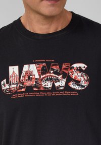 Schwarzes Baumwoll-T-Shirt mit einem großen roten Grafikdesign, das "JAWS" sowie ein Motiv eines Hais, eines Bootes und einer Strandszene zeigt, mit weißem Text darunter.