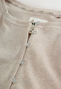 Maglia a coste color beige chiaro con scollo rotondo, cinque bottoni di chiusura e un'etichetta con la scritta "self." stampata all'interno del colletto.