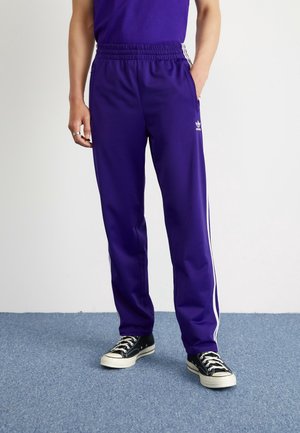 Persoon draagt paarse Adidas trainingsbroek met witte strepen, zwart-witte sneakers, staat op blauw tapijt tegen een witte muur.