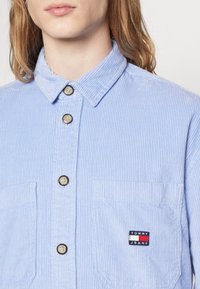 Jasnoniebieska koszula sztruksowa z zapięciem na guziki z przodu, kołnierzykiem w szpic, dwoma przednimi kieszeniami oraz małą naszywką z logo Tommy Jeans na piersi.