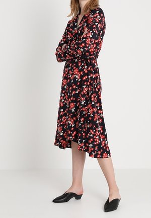 Robe florale noire avec des fleurs rouges et blanches, manches longues, design à nouer, longueur au genou, associée à des chaussures plates noires.