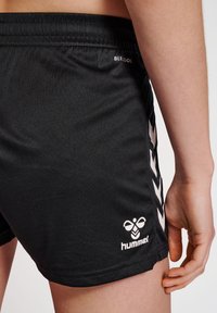 Svart träningsoverallshorts med en slät textur, elastiskt midjeband och en vit logotyp samt chevronmönster på sidan.