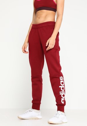 Vrouw draagt rode Adidas sweatpants en een bijpassende sport-bh, gecombineerd met witte sportschoenen, staand tegen een effen achtergrond.