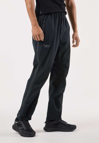 Svarta vattentäta byxor med elastisk midja, utrustade med en dragkedjeförsedd sidficka och reflekterande detaljer för ökad synlighet. Texturerad tygfinish.