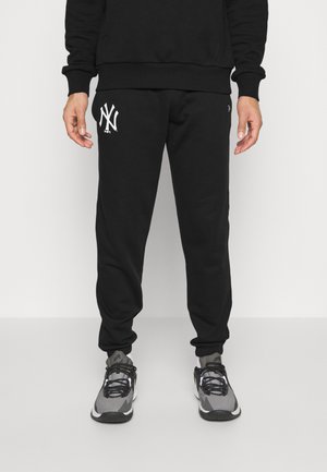 Mężczyzna w czarnych spodniach dresowych z białym logo New York Yankees na lewym udzie i szarymi butami sportowymi, stojący na jednolitym tle.