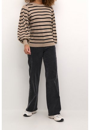 Osoba v béžovo-černém pruhovaném svetru s dlouhým rukávem, černých kalhotách s širokými nohavicemi a bílých teniskách stojící na bílém pozadí.