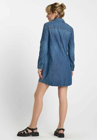 Robe chemise en denim à manches longues, col classique et poignets à boutons. Présente une coupe droite et tombe au-dessus du genou. Portée avec des sandales noires.