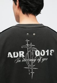 Μαύρο φούτερ με εκτυπωμένη ετικέτα «ADR 0018» και «Στη μνήμη σου» σε λευκό. Περιλαμβάνει διακοσμητικά στοιχεία στο πίσω μέρος.