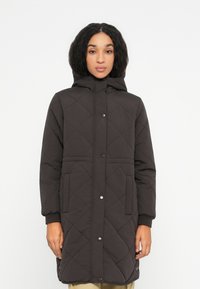 Kvinde iført en lang, sort quilted hættefrakke med trykknapper og lommer, stående mod en plain hvid baggrund.