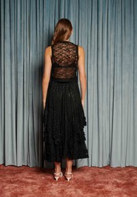 Žena stojaca chrbtom ku kamere, oblečená v čiernom čipkovanom topánke bez rukávov a čiernej vrstvenej sukni s bielymi podpätkami, na pozadí modrých závesov.