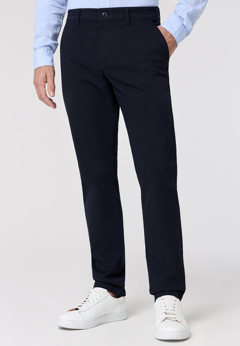 Chino blu navali realizzati in cotone tessuto, con una vestibilità dritta, tasche laterali e una chiusura a bottoni classica, indossati con sneakers bianche.