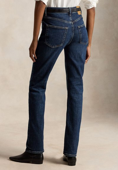 Persona in piedi che indossa jeans blu scuro a gamba dritta, scarpe nere e una camicia bianca a maniche lunghe con le maniche rimboccate.