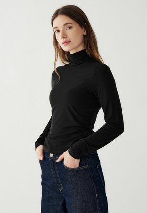 Črn turtleneck dolga majica z dolgimi rokavi, izdelana iz mehkega materiala, prilagojena zasnova z gladko teksturo in visokim ovratnikom. Ujemajo se z temno denim hlačami.