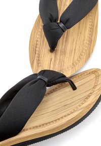 Des brides de thong en tissu noir sur une semelle en bois avec un motif texturé. Le talon est recouvert d'une couche en caoutchouc noir pour l'adhérence et la durabilité.