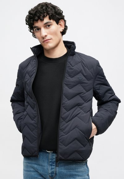 Tmavomodrá prešívaná bunda s vysokým golierom a predným zipsom, s jemným logom a elastickými manžetami, nosená cez čiernu košeľu a rifle.