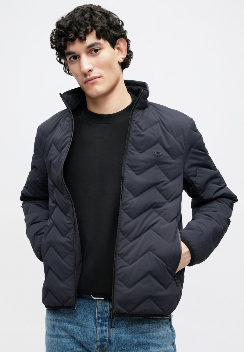 Veste matelassée bleu marine à col montant et fermeture éclair frontale, arborant un logo discret et des poignets élastiques, portée sur une chemise noire et un jean.