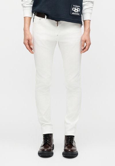 Personne portant un jean blanc slim, une ceinture noire, des bottes montantes en cuir marron foncé et un sweat-shirt bleu marine avec des manches blanches.