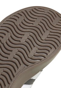 Brązowa podeszwa buta z teksturowanymi falistymi rowkami i prostokątnymi wypustkami, ukazująca część białej i czarnej skórzanej cholewki.