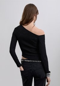 Musta pitkähihainen paita, jossa epäsymmetrinen olkapäästä avoin design ja teksturoitu kangas, yhdistetty korkeavyötäröisiin mustiin farkkuihin ja niitillä koristeltuun vyöhön.