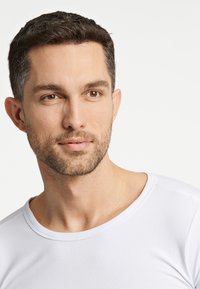 Biała, krótki rękaw T-shirt z okrągłym dekoltem, gładką fakturą i dopasowanym krojem, modelowany przez mężczyznę z krótkimi włosami i brodą.